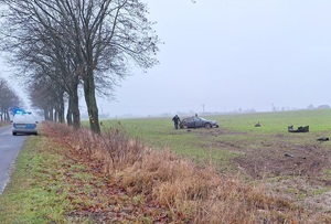 uszkodzony samochód stoja na polu, dwie osoby obok, radiowóz stoi na jezdni